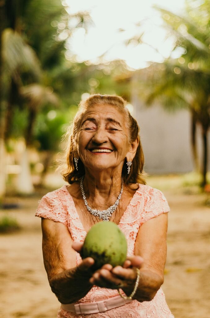 Smiling elderly woman with jewelry showcases happiness while holding a mango outdoors.